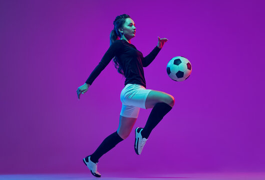 Dynamic portrait of female soccer player practicing with football ball isolated on purple studio background in neon light. Sport, action, motion, fitness