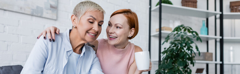 happy woman with cup of tea talking to lesbian girlfriend at home, banner