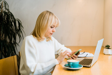 Mature adult woman sitting in cafe with coffee mug and working online on laptop computer. Businesswoman texting messages at mobile phone in coworking space in roasters coffee shop.