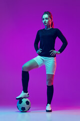 Portrait of beautiful girl, professional female soccer player posing with football ball isolated on purple studio background in neon light. Sport, team, games, fitness