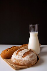 A baguette and a round bread lie on a table covered with a towel, along with a jug of milk. The photo was taken on a black background