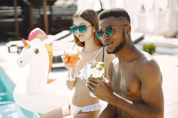 Caucasian girl and black boy sitting near the pool and drinking cocktails