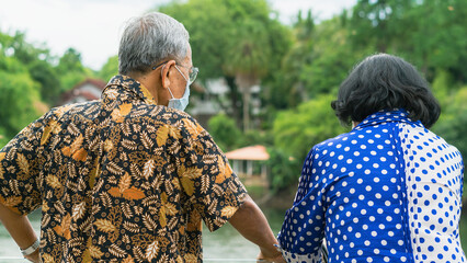 Back view of elderly couple wear protective face mask due to coronavirus (Covid-19) standing side by side at riverside. Relaxed lifestyle for two retired people enjoying freedom in summer holidays.