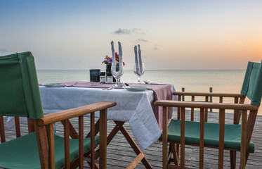 Table set for two on sea coast. Romantic table setting on the sunset. Outdoor restaurant on the beach. Elegant cafe decoration on terrace. Tropical restaurant in summer twilight.