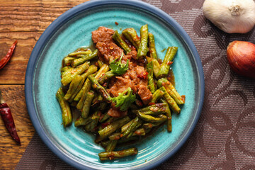 Stir fried pork belly and red curry paste with sting bean served in a blue plate on a wooden table.