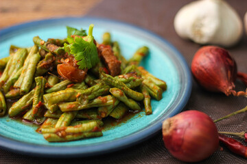 Stir fried pork belly and red curry paste with sting bean served in a blue plate on a wooden table.