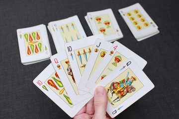 A hand holding a deck of Spanish-suited cards. In the background, more cards from the deck are shown. Colors: black, white and yellow.