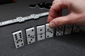Domino tiles placed in a row while a hand places and watches them. In the background, a game of dominoes is being played in a casino.