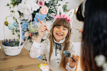Cute little girl in bunny ears with her mother paint eggs while sitting at the table in the kitchen. Decorations for the celebration of Easter