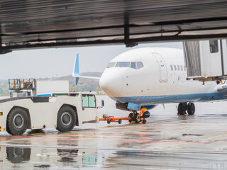 A man in uniform attaches an airfield tractor to an airliner on a rainy day. Aircraft towing