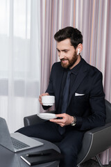 cheerful bearded man in earphone holding cup and looking at laptop