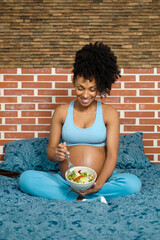 Cheerful black pregnant woman eating healthy salad as snack in her bed. Comfortable expecting mother at home.