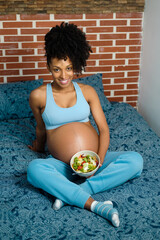 Happy young black pregnant woman holding an organic helathy salad bowl in her bed.