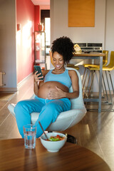 Pregnant woman at home using phone for video call and taking photos to her belly while eating healthy salad. Happy black expecting mother wearing comfortable clothes.