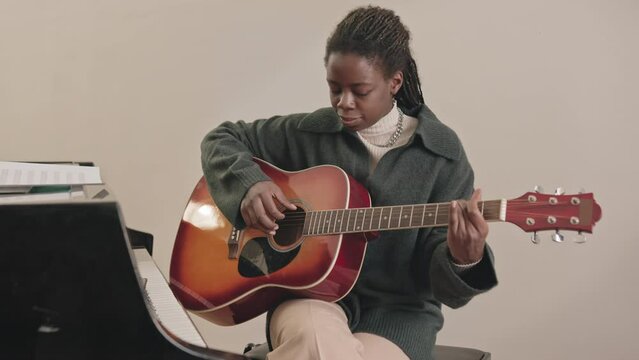 Medium slowmo of young Black woman playing guitar and recording her play on smartphone lying on piano keyboard nearby