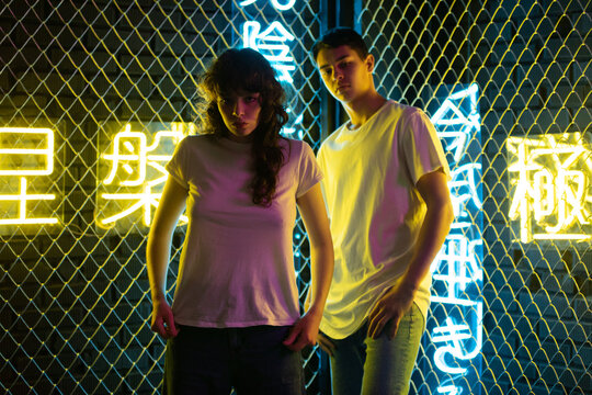 Young couple in white t-shirts stand against the background of a metal grid with neon symbols.