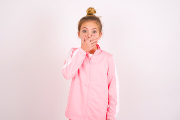 Emotional caucasian little kid girl with bun hairstyle wearing pink tracksuit over white background gasps from astonishment, covers opened mouth with palm, looks shocked at camera. Reaction concept