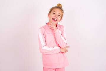Optimistic caucasian little kid girl with bun hairstyle wearing pink tracksuit over white wall keeps hands partly crossed and hand under chin, looks at camera with pleasure. Happy emotions concept.