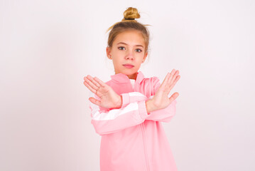 caucasian little kid girl with bun hairstyle wearing pink tracksuit over white background has rejection expression crossing arms and palms doing negative sign, angry face.