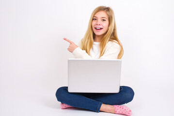 Naklejka premium caucasian teen girl sitting with laptop in lotus position on white background pointing away and smiling to you. Look over there!