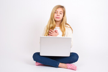 caucasian teen girl sitting with laptop in lotus position on white background closes eyes and keeps hands on chest near heart, expresses sincere emotions, being kind hearted and honest.