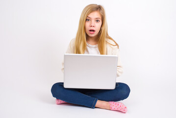 caucasian teen girl sitting with laptop in lotus position on white background having stunned and shocked look, with mouth open and jaw dropped exclaiming: Wow, I can't believe this. Surprise and shock