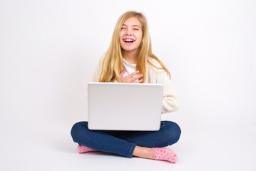 Fototapeta premium caucasian teen girl sitting with laptop in lotus position on white background keeps hands crossed, laughs out loud at good joke, wears casual clothes.