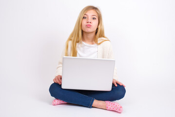 caucasian teen girl sitting with laptop in lotus position on white background , keeps lips as going to kiss someone, has glad expression, grimace face. Standing indoors. Beauty concept.