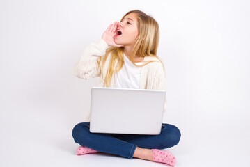 Fototapeta premium caucasian teen girl sitting with laptop in lotus position on white background profile view, looking happy and excited, shouting and calling to copy space.