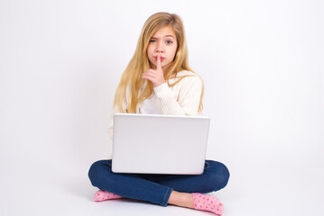 Naklejka premium caucasian teen girl sitting with laptop in lotus position on white background makes hush gesture, asks be quiet. Don't tell my secret or not speak too loud, please!