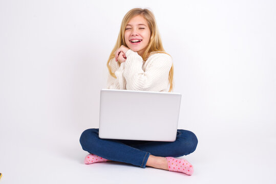Dreamy caucasian teen girl sitting with laptop in lotus position on white background with pleasant expression, closes eyes, keeps hands crossed near face, thinks about something pleasant