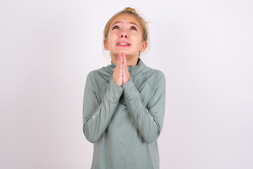 little caucasian kid girl with hair bun wearing technical shirt over white background begging and praying with hands together with hope expression on face very emotional and worried. Please God
