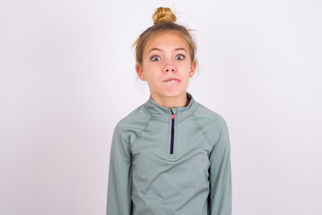 little caucasian kid girl with hair bun wearing technical shirt over white background making grimace and crazy face, screaming out of control, funny lunatic expressing freedom and wild.