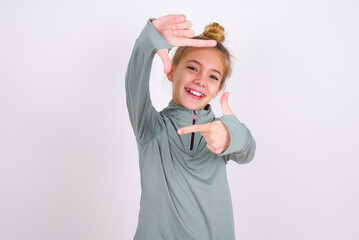 little caucasian kid girl with hair bun wearing technical shirt over white background making finger frame with hands. Creativity and photography concept.