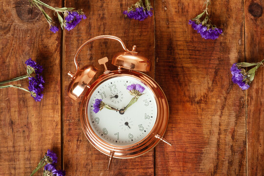 Vintage, beautiful alarm clock made from gold metal with blue flowers instead of clock's hands and flowers around it on wooding surface. top view. Spring time. Time for spring. Conceptual pic