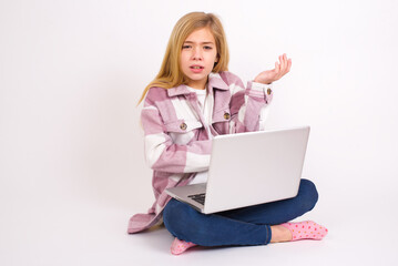 Studio shot of frustrated caucasian teen girl sitting with laptop in lotus position on white background gesturing with raised palm, frowning, being displeased and confused with dumb question.