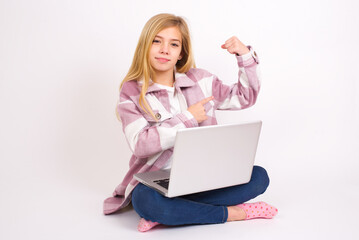 Fototapeta premium Smiling caucasian teen girl sitting with laptop in lotus position on white background raises hand to show muscles, feels confident in victory, strong and independent.
