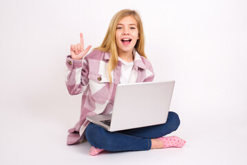 Pleasant looking caucasian teen girl sitting with laptop in lotus position on white background has clever expression, raises one finger, remembers himself not to forget tell important thing.