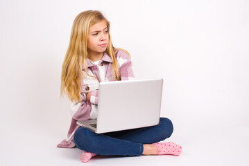 Displeased caucasian teen girl sitting with laptop in lotus position on white background with bad attitude, arms crossed looking sideways. Negative human emotion facial expression feelings.