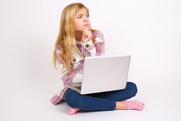 caucasian teen girl sitting with laptop in lotus position on white background with hand under chin and looking sideways with doubtful and skeptical expression, suspect and doubt.