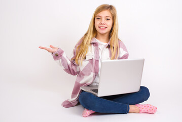 Positive glad caucasian teen girl sitting with laptop in lotus position says: wow how exciting it is, has amazed expression, shows something on blank space with open hand. Advertisement concept.