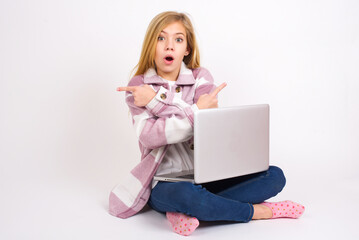 Confused caucasian teen girl sitting with laptop in lotus position on white background chooses between two ways, points at both sides with crossed hands, feels doubt. Need your advice.