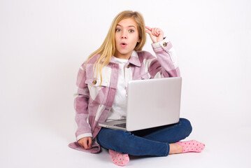 Naklejka premium Shocked caucasian teen girl sitting with laptop in lotus position on white background shows something little with hands, demonstrates size, opens mouth from surprise. Measurement concept.