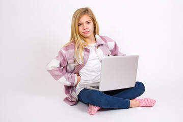 Funny frustrated caucasian teen girl sitting with laptop in lotus position on white background holding hands on waist and silly looking at awkward situation.