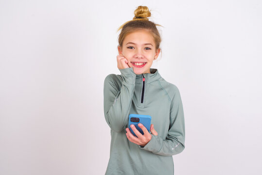 Joyous little caucasian kid girl with hair bun wearing technical shirt over white wall poses with mobile phone device, types text message on modern smartphone, watches funny video during free time,