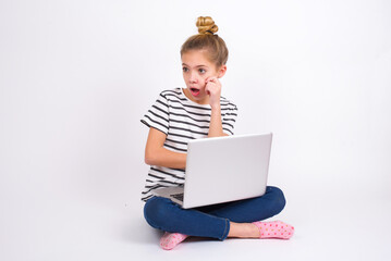 Astonished caucasian teen girl sitting with laptop in lotus position on white background looks aside surprisingly with opened mouth.