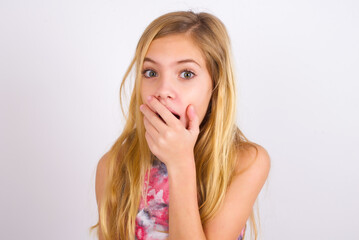 little caucasian kid girl wearing sport clothing over white background covers mouth and looks with wonder at camera, cannot believe unexpected rumors.