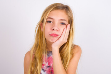 Sad lonely little caucasian kid girl wearing sport clothing over white background touches cheek with hand bites lower lip and gazes with displeasure. Bad emotions
