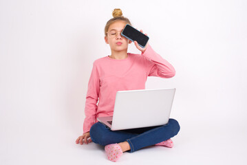 Adorable Beautiful caucasian teen girl sitting with laptop in lotus position on white background holding modern device covering eye with lips pouted