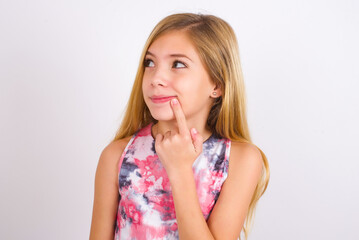 Lovely dreamy little caucasian kid girl wearing sport clothing over white background keeps finger near lips looks aside copy space.